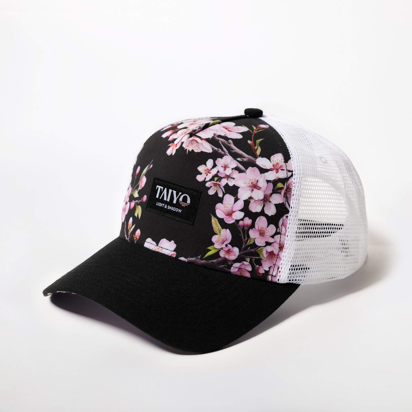 Casquette Sakura Trucker Black de TAIYO avec imprimé fleurs de cerisier sur le panneau avant et sous la visière, filet arrière blanc et patch tissé noir.